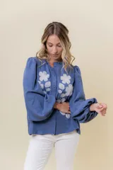 Camisa azul de calce relajado con bordado floral blanco en el frente. Presenta mangas largas amplias con ligero fruncido en el hombro y cierre frontal con botones.