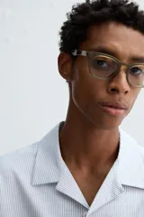 Camisa de algodón con rayas verticales finas azules y blancas, corte relajado (relaxed fit), cuello bowling y manga corta. Posee un bolsillo plastrón en el pecho y aberturas laterales en el bajo.