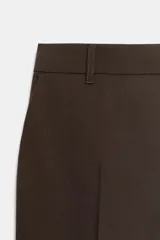 Pantalón de corte recto y tiro bajo, color marrón oscuro. Presenta trabillas en la cintura, bolsillos laterales, falsos bolsillos traseros de vivo y raya planchada en el delantero. Cierre frontal con cremallera, botón interior y ganchos metálicos.