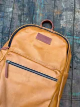 Mochila de cuero color tostado con bolsillo frontal con cierre, asa de mano superior y etiqueta de cuero con la marca grabada.
