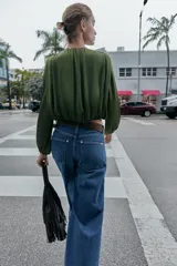 Blusa de manga larga color verde oliva, confeccionada en tejido satinado con textura acanalada. Presenta escote redondo con lazo ajustable que termina en un dije dorado, y el bajo es abullonado con elástico interior.