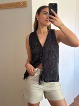 Chaleco tejido en punto calado con hilo de algodón y lurex, de color negro con destellos plateados. Presenta escote profundo en V, cierre frontal con cordones entrelazados terminados en borlas y un corte recto y holgado.
