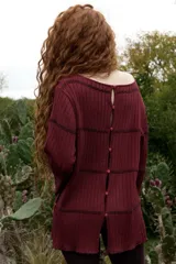 Buzo reversible de tejido fino con sutil brillo, color bordó, con cuello bote y mangas largas acampanadas. Presenta costuras decorativas horizontales y botones en forma de flor a los costados.