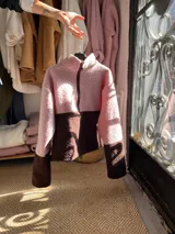 Buzo de polar tipo corderito con diseño de bloques de color en tonos rosa y marrón chocolate. Presenta cuello alto con cierre frontal parcial y un aplique circular en el pecho.