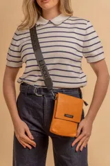 Phonebag pequeña color mostaza, confeccionada en cuero sintético, con solapa frontal y correa ajustable con logo de la marca.