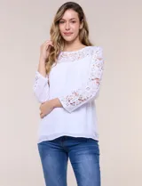 Blusa blanca de gasa liviana con forro interno en el cuerpo, de estilo boho-chic y silueta semi holgada. Presenta escote redondo con canesú de encaje calado en el frente y mangas largas con paneles longitudinales de encaje.