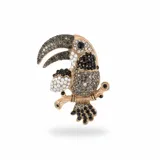 Broche con forma de tucán posado sobre una rama, elaborado en metal dorado con incrustaciones de strass en tonos negro, gris y plateado.