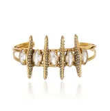 Pulsera rígida tipo brazalete de metal dorado con acabado envejecido, presenta un diseño con estructuras verticales texturizadas con pequeños cristales y piedras centrales transparentes en forma de marquise.