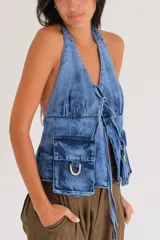 Top halter de denim azul con efecto deslavado, escote pronunciado, cierre frontal con tiras para anudar, bolsillos cargo delanteros con detalles de estribos metálicos y diseño utilitario.