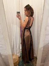 Vestido largo de seda color bronce con acabado brillante, escote profundo en V y espalda abierta. Presenta detalles en terciopelo negro que enmarcan el escote, recorren el frente y definen la cintura con una faja ajustable. Posee breteles anchos y una abertura frontal en la falda.