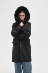 Campera parka negra de corte largo y holgado, con capucha fija adornada con piel sintética, cierre frontal oculto y bolsillos laterales con cordones ajustables en la cintura.