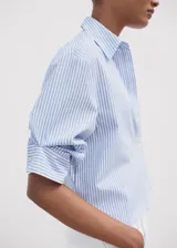 Camisa de corte crop y diseño recto, con estampado de rayas verticales en azul y blanco. Presenta cuello camisero, bolsillo en el pecho, cierre frontal con botones y mangas tres cuartos.
