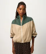 Campera rompeviento de diseño tricolor con bloques de color en verde, beige y blanco. Presenta cuello alto, cierre frontal con cremallera, mangas raglán y puños ajustados. El ruedo incluye un cordón regulable para un ajuste personalizado.
