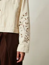 Campera de denim color off white con cuello camisero, bolsillos con solapa en el pecho y cierre frontal con botones metálicos. Presenta detalles de bordado con perforaciones en las mangas.