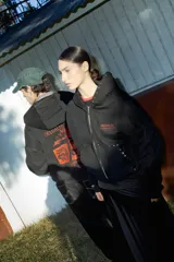 Campera negra de algodón con cierre frontal, capucha, bolsillos laterales y detalles de tachas metálicas en la capucha y los bolsillos. Presenta un estampado frontal con texto en tono rojizo.