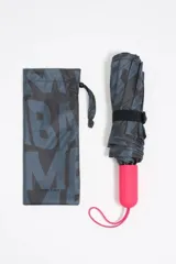 Paraguas plegable con funda textil estampada con el logo de Bimba y Lola en tonos oscuros (negro/gris). El mango es de goma en color fucsia vibrante con el nombre de la marca grabado.