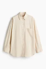 Camisa oversized de color beige con diseño de rayas finas, cuello clásico, cierre frontal de botones, bolsillo en el pecho y mangas largas con puños abotonados.