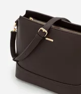 Cartera tipo hobo de tamaño pequeño, confeccionada en material sintético color marrón oscuro con acabado liso. Presenta una correa ajustable, cierre principal con cremallera y detalles metálicos dorados, incluyendo una placa con el logo de la marca en el frente.