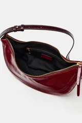 Bolso de hombro de diseño semicircular, confeccionado en material sintético efecto charol de color rojo. Cuenta con una correa ajustable fina con detalles metálicos dorados y cierre superior con cremallera.