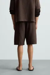 Bermuda de corte regular fit, color marrón oscuro, confeccionada en tejido con lino. Presenta pliegues frontales, bolsillos delanteros y bolsillos traseros de vivo. Cierre frontal con cremallera y botón.