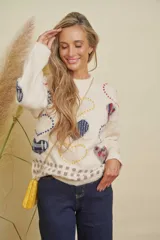 Buzo de punto color crema con estampado de corazones en tonos azul, rojo y amarillo, y detalles cuadrados en gris en el ruedo. Tiene cuello redondo y mangas largas.