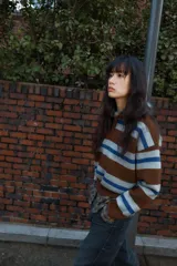 Jersey de punto con diseño a rayas horizontales en tonos marrón, gris y azul. Presenta cuello tipo polo con abertura frontal, manga larga y puños acanalados.