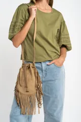 Cartera tipo bucket bag color camel, confeccionada en material sintético con acabado gamuzado. Presenta un diseño decorado con flecos largos en todo el contorno, cierre mediante cordón ajustable y correa larga regulable.