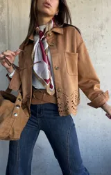 Chaqueta de gamuza color camel con cierre frontal de botones, cuello clásico y detalle de calados decorativos en el ruedo. Presenta bolsillos frontales y mangas largas con puños.