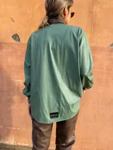 Camisa verde de cuero sintético con cuello marrón, cierre frontal con botones, dos bolsillos con solapa en el pecho y cinturón marrón.