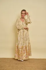 Vestido largo dorado con estampado floral y detalles metalizados, escote en V, mangas largas con puños ajustables con cordones y volados en la falda.