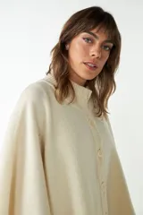 Capa color crudo de tejido de punto 100% lana fina, con cuello alto, cierre frontal con botones, bolsillos delanteros y corte asimétrico.