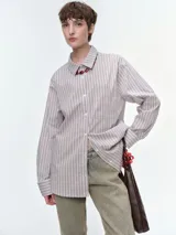 Camisa oversize de popelina de algodón con diseño de rayas verticales en tonos rosado y marrón. Presenta cuello clásico, cierre frontal con botones, mangas largas con puños abotonados y un bolsillo aplicado en el pecho.