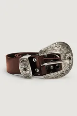 Cinto de cuero marrón con gran hebilla metálica plateada de estilo western, con grabados ornamentales, detalles de tachas y apliques circulares oscuros.