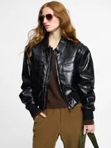 Chaqueta biker corta negra de cuero sintético, con cuello clásico, cierre frontal con cremallera, bolsillos con solapa y puños ajustados.