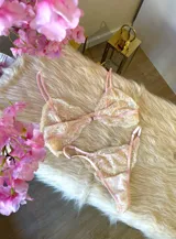 Conjunto de lencería compuesto por un bralette y una tanga, confeccionados en encaje bitono rosa y champagne con diseño floral. El bralette presenta cierre frontal con broche de gancho y breteles regulables. La tanga es de estilo colaless con laterales regulables.