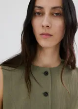 Vestido corto sin mangas color verde militar, con cuello redondo y cierre frontal con botones oscuros. Presenta bolsillos de ribete en la parte delantera y un diseño de corte recto.