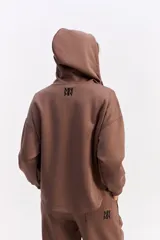 Hoodie de algodón color marrón con capucha, cierre frontal parcial y detalle de costuras horizontales en contraste negro. Presenta diseño holgado, mangas largas con puños elásticos y cordones ajustables en la capucha.