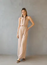 Mono largo de color nude, con escote halter y detalle de cadena en el cuello. Presenta una silueta relajada con pantalón de corte recto y fluido.