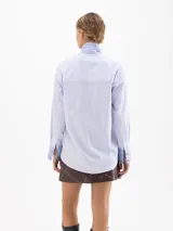 Camisa de poplín con diseño de rayas verticales en tonos azul y blanco. Presenta un cuello prolongado que se anuda en forma de lazo frontal, mangas largas con puños anchos y corte holgado.