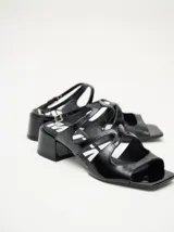 Sandalias de cuero charol negro con múltiples tiras cruzadas en el empeine, punta cuadrada y taco bloque de altura media. Cuenta con correa ajustable en el tobillo con hebilla metálica.