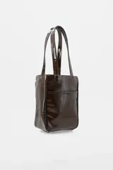 Bolso tipo tote de charol color marrón, con diseño vertical, doble asa de hombro y logo de la marca estampado en la parte inferior. Cuenta con terminaciones en contraste y estructura rígida.