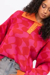 Buzo de tejido de punto con diseño geométrico en tonos fucsia y rojo, cuello tipo polo con cierre de botones y puños en color naranja contrastante. Presenta un corte holgado y oversize.