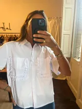 Camisa blanca de manga corta con cuello clásico y cierre de botones. Presenta dos bolsillos frontales con volantes decorativos.