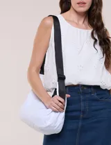 Cartera bandolera de lona blanca con silueta de medialuna, cierre superior negro y correa regulable en contraste.