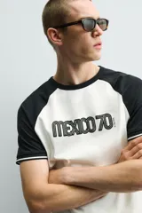 Camiseta de corte regular con diseño color block, cuerpo en color blanco y mangas ranglán en color negro. Presenta cuello redondo, detalles de rib a contraste en los puños y estampado frontal con la leyenda Mexico 70.