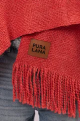 Ruana de tejido de punto color coral con flecos en el ruedo y etiqueta de cuero con logo grabado en la parte frontal.