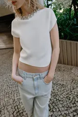 Top blanco de punto con cuello redondo adornado con una aplicación de bolas plateadas, manga corta y bajo asimétrico. Cuenta con una abertura trasera con botón oculto.