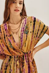 Vestido corto de estilo kimono con estampado tie-dye en tonos mostaza, violeta y negro. Presenta escote en V profundo, mangas cortas amplias y ajuste en la cintura con cordón. Incluye detalles de bordados decorativos con cuentas en el escote y la falda.