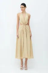 Vestido midi de tricolina en color beige miel, con cuello halter, falda amplia y diseño sin mangas. Presenta un detalle de giro y lazo en la parte delantera, con aberturas laterales en la cintura y cierre de cremallera invisible en la espalda.