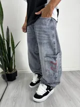 Pantalón de jean estilo baggy con lavado ácido en tono azul grisáceo, corte holgado y diseño de bolsillos tipo cargo con detalles bordados en rojo.